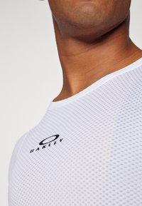 T-shirt de sport en mesh blanc avec un col rond et des manches courtes, arborant un logo noir Oakley sur le devant. Texture légère et respirante.