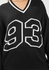 Mustassa neulotussa villapuserossa on V-pääntie, ja sen edessä on rohkeat valkoiset numerot "93" ja "3" suurena, geometrisena muotona.