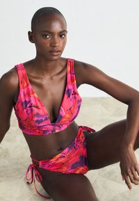 Cvetni bikini komplet v rožnatih in vijoličnih tonih, ki vključuje globok zgornji del s širokimi naramnicami in nastavljivimi stranskimi vezalkami, izdelan iz gladkega materiala.