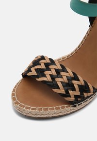 Gros plan de l'avant d'une sandale avec une lanière en cuir tressé beige et noir et une semelle marron clair avec des coutures visibles sur fond blanc.