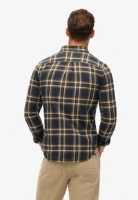 Camicia di flanella a quadri in blu navy, crema e rosso con maniche lunghe, colletto con bottoni e spalla raglan. Caratterizzata da una vestibilità comoda e una texture sottile.