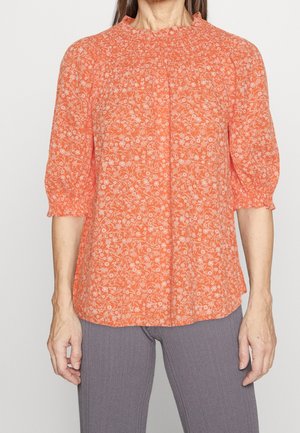 Kobieta w koralowej bluzce z białym kwiatowym wzorem i rękawami 3/4 z bufkami, zestawiona z szarymi prążkowanymi spodniami.