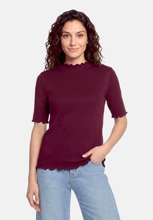 Femme aux cheveux bouclés foncés portant un haut bordeaux à manches courtes avec des bords festonnés et un jean bleu clair, debout avec une main dans la poche.
