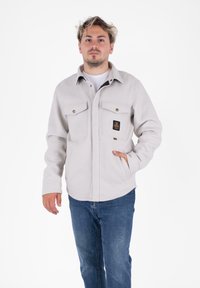 Giacca camicia in flanella grigia chiara con tasche sul petto con bottoni, tasche laterali e un'etichetta nera sul lato sinistro. Texture morbida e vestibilità rilassata.