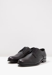 Paire de chaussures formelles noires en cuir à lacets sur une surface blanche avec un fond gris clair uni.