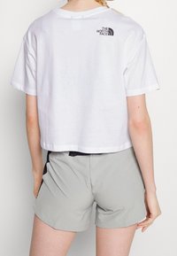 Bílý zkrácený tričko z bavlny, s malým černým logem na horní části zad, v kombinaci se světlými šedými šortkami.
