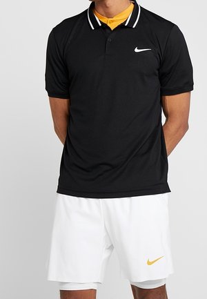 Homme portant un polo noir Nike avec des rayures blanches au col, un sous-vêtement jaune, et un short blanc Nike avec un logo jaune, debout sur un fond gris.