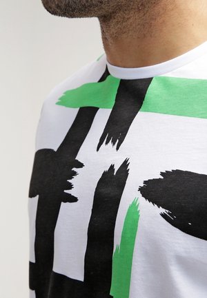 Witte T-shirt met een gedurfde abstracte print bestaande uit grote zwarte en groene penseelstreken. Zacht katoen materiaal, ronde halsstijl.