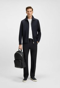 Men's outfit features a black zip-up jacket, white t-shirt, black tailored pants, and a textured black leather backpack. Sneakers with light soles.