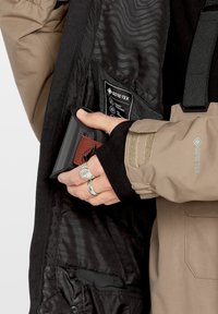 GORE-TEX jas met een zwarte binnenkant en een beige buitenkant; heeft een gladde, waterdichte stof, een zak en een gestructureerde voering. Zilveren ring zichtbaar.