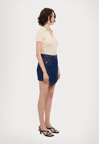 Camisa polo de punto beige con mangas cortas, combinada con una falda envolvente de mezclilla azul. La modelo lleva sandalias negras de tiras. El fondo es blanco.