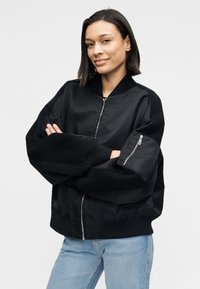 Μαύρο μπουφάν βομβιστή από λείο ύφασμα με φαρδιά μανίκια, ριμπ μανσέτες, φερμουάρ μπροστά και πλαϊνή τσέπη με φερμουάρ. Συνδυασμένο με ανοιχτόχρωμα μπλε τζιν.