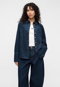 Σκούρο πουκάμισο από τζιν με κουμπιά, γιακά, μακριά μανίκια και κλείσιμο με κουμπιά μπροστά. Φορεμένο πάνω από ένα λευκό μπλουζάκι, συνοδευόμενο από casual παντελόνια με ευθείες γραμμές.