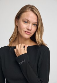 Maglietta nera a maniche lunghe con coste, scollo ampio e dettagli ricamati sui polsini, indossata da una donna con capelli mossi.