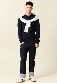 Maglione blu navy a maglia con dettagli a coste, sciarpa bianca drappeggiata attorno al collo, jeans in denim scuro con orli arrotolati e sneakers bianche con dettagli neri.