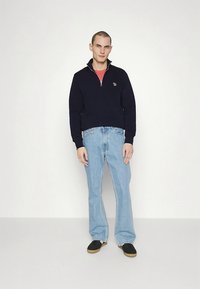 Tengerészkék cipzáras pulóver álló gallérral, világoskék bővülő farmerral és fekete cipővel párosítva. A mellrészen hímzett logóval.