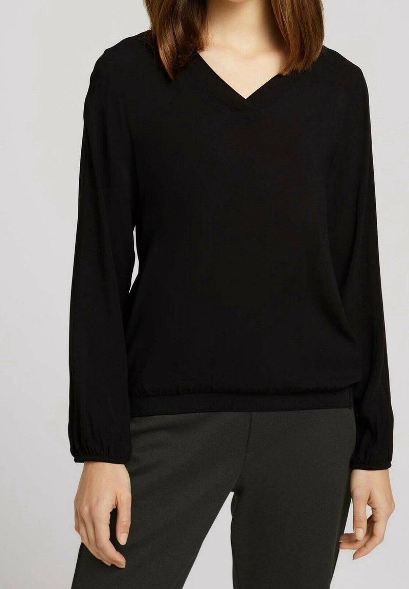 Blusa nera a maniche lunghe con scollatura a V, caratterizzata da una vestibilità ampia e dettagli con polsini arricciati. Abb bins con pantaloni grigio scuro.