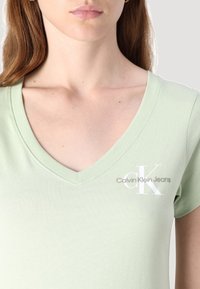 Svetlozelené tričko s výstrihom do V vyrobené z jemnej textílie, s bielym logom "CK" a textom "Calvin Klein Jeans" v sivej farbe na ľavej strane hrude.