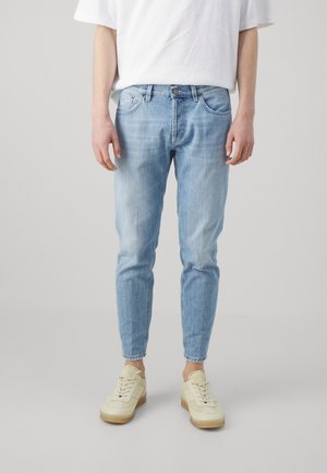 Person trägt hellblaue Slim-Fit-Jeans, weißes T-Shirt und cremefarbene Sneakers und steht vor einem schlichten Hintergrund.