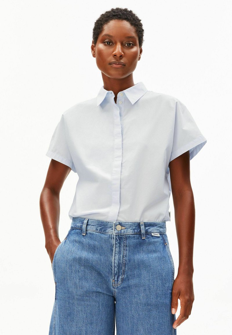 Femme aux cheveux courts et bouclés portant une chemise bleu clair à manches courtes avec col et un jean bleu taille haute, debout les mains dans les poches.