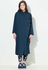Abito avvolgente teal con maniche lunghe e dettagli con bottoni, abbinato a leggings con motivo galattico e sneakers blu scuro. Tessuto morbido.