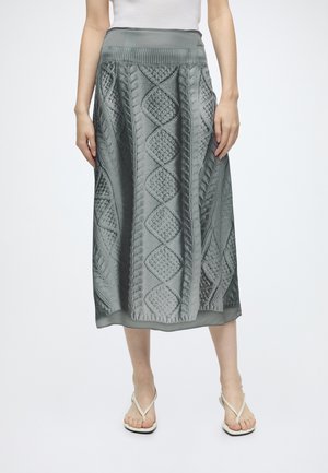 Femme portant une jupe midi en tricot gris avec des motifs texturés en losange et câble, associée à des sandales blanches à brides et un haut blanc sans manches.