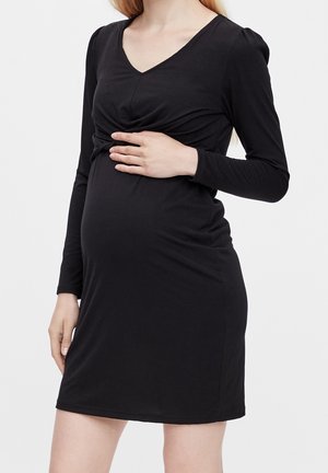Kobieta w ciąży w dopasowanej czarnej sukience z długim rękawem, delikatnie trzymająca brzuch jedną ręką.