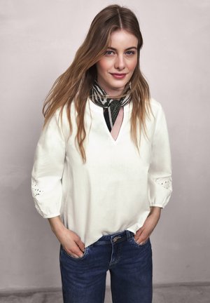 Młoda kobieta z długimi włosami, ubrana w białą bluzkę, wzorzysty szalik na szyi oraz niebieskie dżinsy, stoi z rękami w kieszeniach na jednolitym tle.