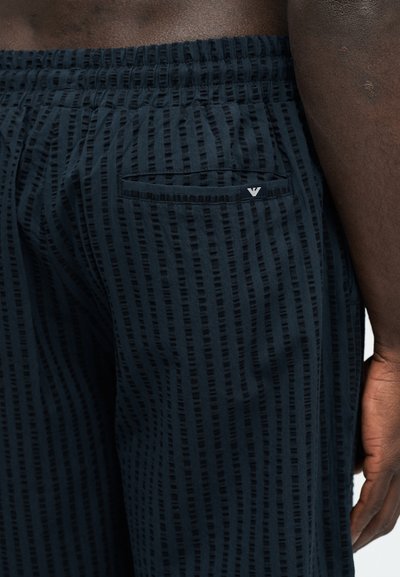 Pantalones cortos de textura azul oscuro con rayas verticales y un pequeño logotipo en el bolsillo trasero, usados por una persona de piel oscura con una mano visible.
