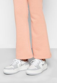 Osoba w białych i jasnoszarych sneakersach, ubrana w jasnoróżowe, szerokie spodnie, stojąca na szarej powierzchni na tle jednolitego tła.