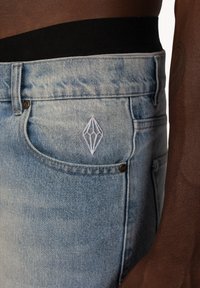 Jasnoniebieskie dżinsy mają klasyczny pięciokieszeniowy krój, z białym haftowanym logo w kształcie diamentu na przedniej kieszeni oraz czarnym pasem.