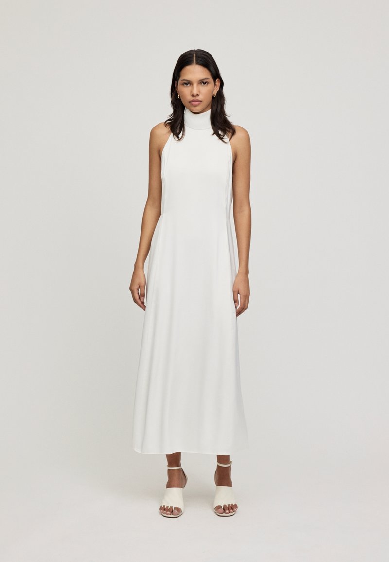 Vestido largo sin mangas de color blanco con un escote alto, textura suave y un corte holgado; combinado con sandalias ligeras de punta abierta.