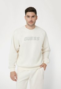 Kreminė palaidinė su apvaliu iškirpimu ir raglan rankovėmis. Puikuojasi iškilus pilkas "GUESS" logotipas ant krūtinės. Minkšta tekstūra, atsipalaidavęs siluetas.