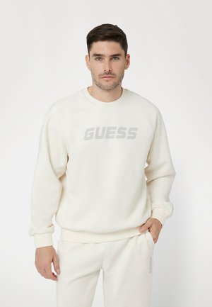 Kreminė palaidinė su apvaliu iškirpimu ir raglan rankovėmis. Puikuojasi iškilus pilkas "GUESS" logotipas ant krūtinės. Minkšta tekstūra, atsipalaidavęs siluetas.