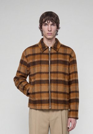 Joven con chaqueta marrón y naranja a cuadros con cremallera y cuello, y pantalones beige, de pie frente a un fondo claro liso.