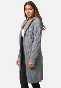Lungo cardigan grigio a maglia con cappuccio. Presenta un motivo a trecce, polsini a costine e una vestibilità rilassata. Indossato sopra una maglietta bianca e pantaloni neri.