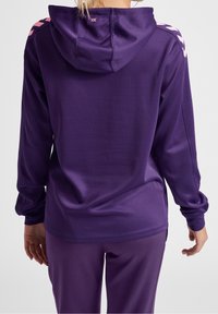 Fialová mikina s měkkou texturou, má kapuci, dlouhé rukávy a růžové akcentové pruhy na ramenou. Vybavená se shodnými kalhotami.