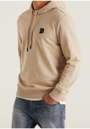 Uomo che indossa un felpa con cappuccio beige con patch logo nero e jeans blu, con una mano in tasca, in piedi contro uno sfondo neutro.