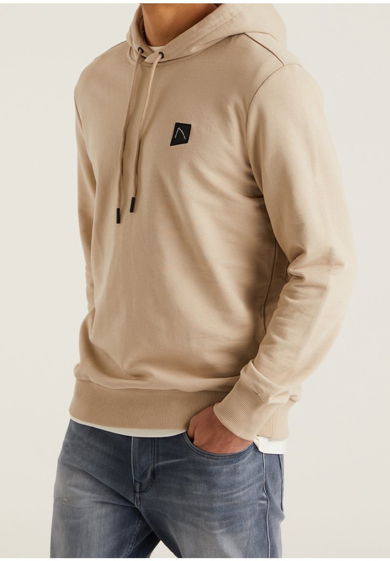 Uomo che indossa un felpa con cappuccio beige con patch logo nero e jeans blu, con una mano in tasca, in piedi contro uno sfondo neutro.