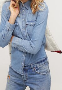 Person som bär en ljusblå denimskjorta och jeans med en liten regnbågsapplikation, håller en vit sherpa-jacka över axeln.