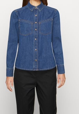 Femme portant une chemise en denim bleue à boutons avec poches poitrine et pantalon noir, debout devant un fond clair uni.