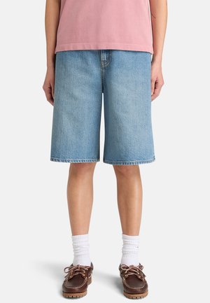 Person wearing light blue denim knee-length shorts, white crew socks, brown leather boat shoes, and a pink shirt, standing against white background.