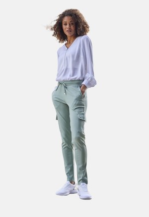 Hellgrüne Cargo-Hosen mit einem Kordelzugbund, Seitentaschen, kombiniert mit einer weißen Langarmbluse und weißen Sneakers.