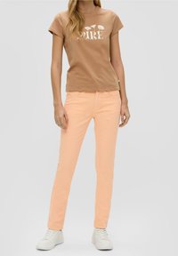 Donna che indossa una maglietta marrone con testo bianco a forma di fiore e pantaloni slim-fit pesca, abbinati a sneaker bianche, in piedi su uno sfondo grigio semplice.