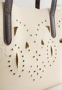Sac fourre-tout crème avec des poignées brun foncé, présentant des motifs découpés complexes de fleurs et de paisley brodés sur le devant.