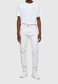 Man wearing white jeans, white high-top sneakers, and a white T-shirt with a small colorful circular logo on the chest.