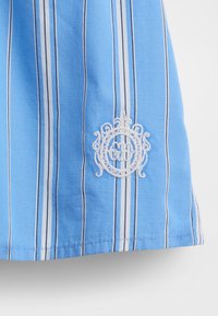 Tela de rayas azules con líneas blancas y oscuras. Presenta un emblema bordado en blanco cerca del dobladillo, que resalta el diseño intrincado.