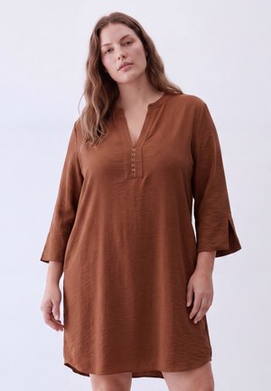 Woman with long wavy hair wearing a loose brown dress with three-quarter sleeves and a buttoned V-neck, standing against a plain light background.