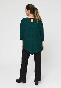 Zelena teksturirana bluzica s tričetvrt rukavima, koja ima detalj u obliku ključanice na leđima. Uparićena s crnim hlačama i robustnim crnim petama.