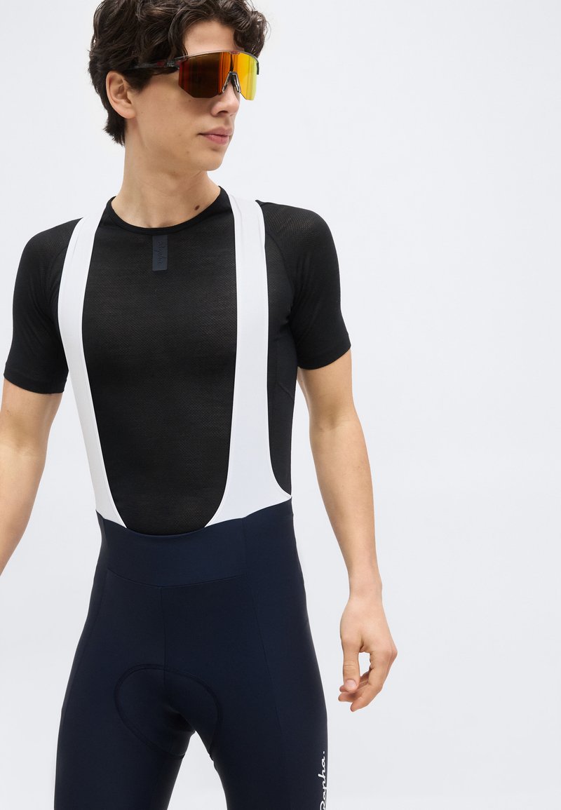 Young man wearing black cycling bib shorts with white straps and a black mesh short-sleeve base layer, looking to the side, wearing orange-tinted sunglasses.